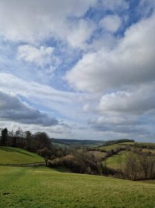View over rolling hills, hedges and fields stretching off into the distance under a blue sky with clouds