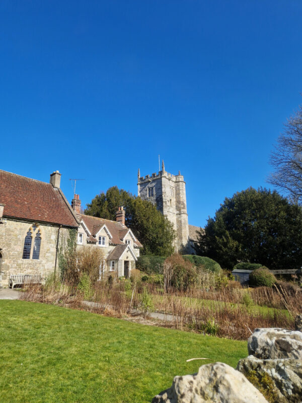 View of a cottage and garden with a church tower in the background against a bright blue sky