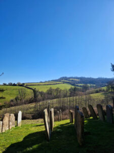 View out over a graveyard with ancient headstones in the foreground and rolling hills stretching off into the distance.