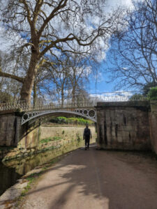 A person walking next to a canal under and ornate Victorian white iron bridge