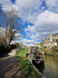 A narrowboat moored on the canal in Bath, with an ornate, white Victorian iron bridge in the backgruond