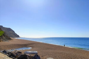 View of a pebble beach looking out to sea with a cliff rising up from the sea at the far left of the beach. The sea is calm, there isn't a cloud in the sky and the sun is shining. A lone figure walks along the beach at the water's edge.