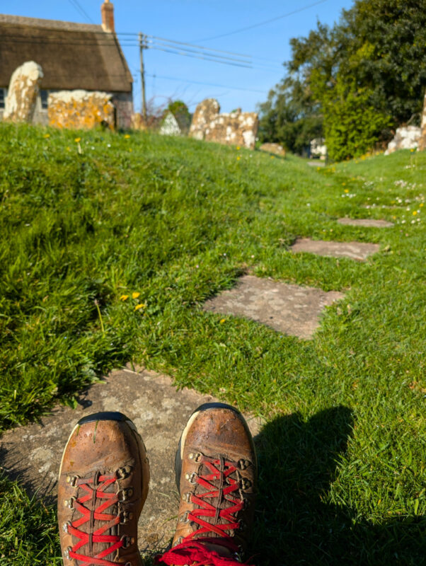 Photo of a pair of walking boots with red laces in front of an old stone path in a graveyard. There are lichen-covered gravestones in the distance.