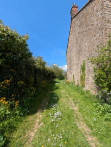 A grass footpath with an old stone building on the right and a hedgerow on the left