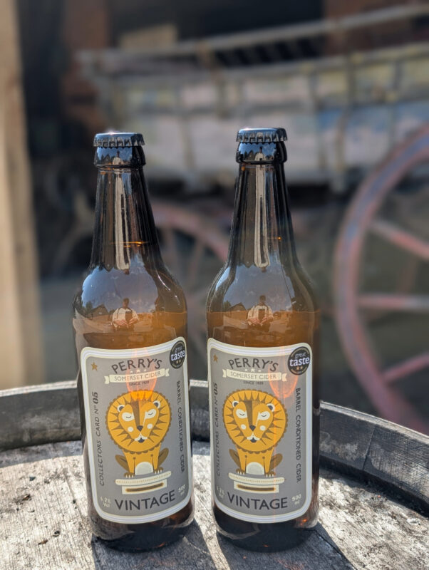 Photo of two bottles of cider on a wooden barrel with an old cart in the background
