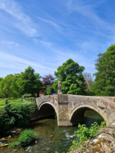 An old stone bridge with two arches and a statue in the middle. The bridge is surrounded by lush green trees,