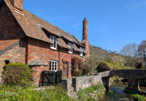 An old red brick cottage with a large chimney next to a bridge going over a river.