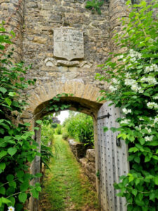 An old stone archway and open door below a family crest. A white rose is growing up the wall to the right.