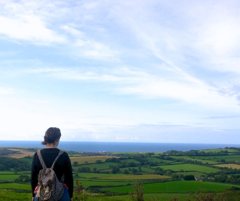 A female hiker stands with her back to the camera looking out over the landscape to the sea