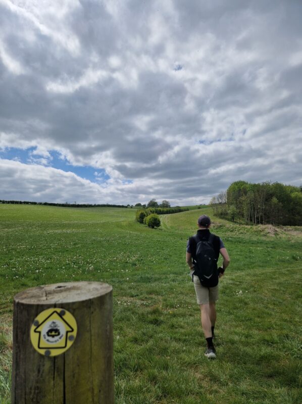 A hiker striding off into the distance across a field with a waymarker in the foreground