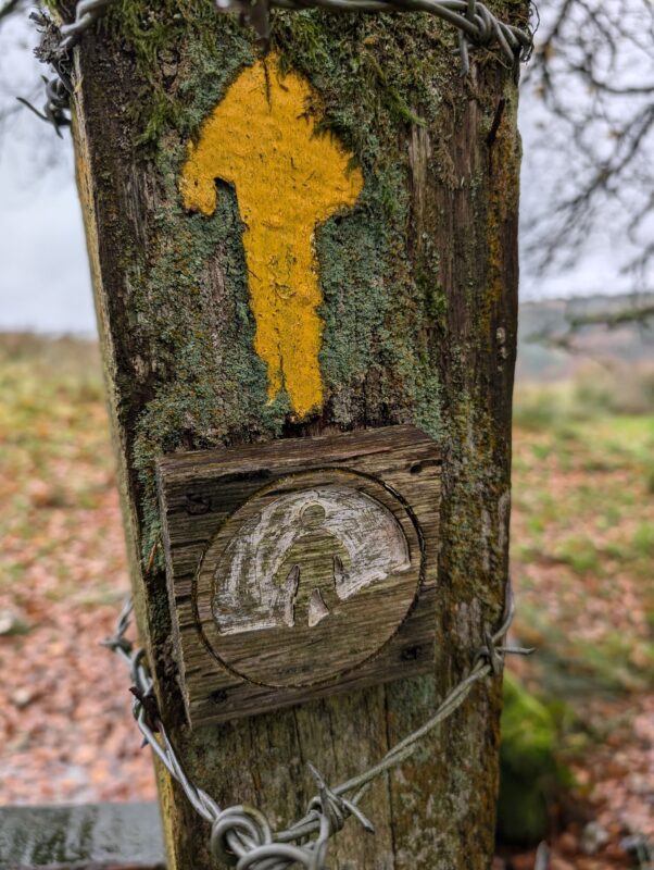 An old wooden waymarker with a yellow arrow and image of a person walking carved into the wood