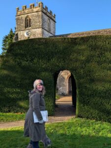 A female walker standing in front of a large sculptured hedge with a church tower peeking out above the hedge in the background