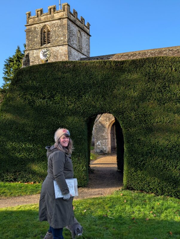 A female walker standing in front of a large sculptured hedge with a church tower peeking out above the hedge in the background