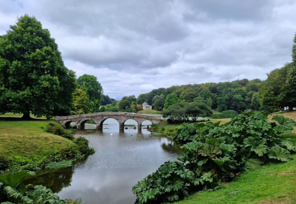 A Palladian bridge over a large landscapes lake with a temple in the background