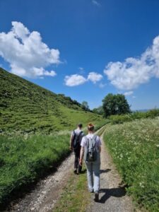 A man and a woman walking down a dirt track with bordered by green fields under a bright blue sky