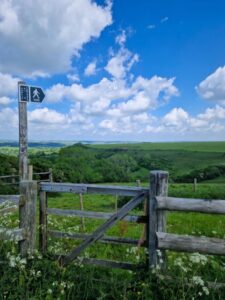 A footpath fingerpost and a gate leading into a field with views stretching off into the distance over rolling green countryside