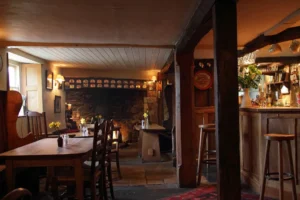 A cosy country inn with wooden beams, a large stone fireplace in back, tables on the left and a bar to the right.