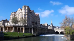 A view of the centre of Bath: Pulteney bridge and weir to the right and the grand Empire hotel with a green in front of it to the left.
