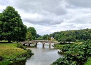 A Palladian bridge over a lake in the landscaped gardens of a country house. A Greek-inspired temple is in the background and there are lush green trees and foliage all around it.