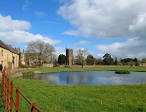 A traditional English village with a pond in the centre, stone cottages around the side and a church tower in the distance