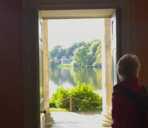 A hiker looking through a doorway onto a lake and a Greek-inspired temple in the landscaped gardens of a country house.