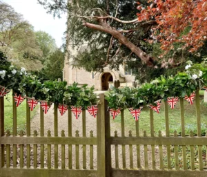 Photo of wooden church gates decorated with green and white leaf and flower garland and union jack flags. The church entrance can be seen in the background partly obscured by trees.