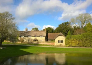 A traditional thatched cottage and outbuildings next to a lake. A small lane passes in front of the cottage with trees and hedges to the right and left of the buildings.
