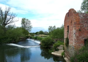 The ruins of an old brick building on the edge of a river with a weir in the background
