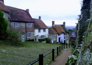 Image looking down an ancient cobbled street with traditional cottages to the left descending down the hill, railings separating a footpath from the road and a stone wall to the right.