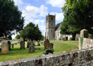 View of a church on a summer's day. There is an old stone wall at the front, behind this a graveyard with the church at the back of the scene.