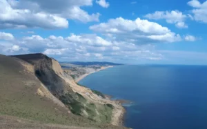 A dramatic shot of the coast in South West England. Steep cliffs on the left side of the photo fall away to the sea in the centre and right. The coast stretches off into the distance under a blue sky with white clouds.