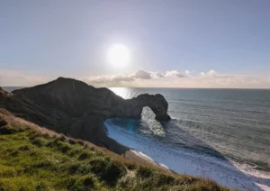 Image of Durdle Door on a sunny day: A natural stone arch jutting out into the sea. The sun behind the arch casts long shadows on the beach. The photo is taken from the South West Coast Path on a cliff looking down on the arch.
