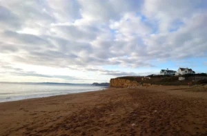 Photo of a sandy beach in the evening with cliffs stretching off into the distance and the sea to the left. A hotel is located on the right on top of a cliff.