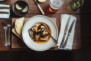 A bowl of mussels on a country inn dining table with cutlery, serviettes, a pint of beer and some lemons in a bowl of water.