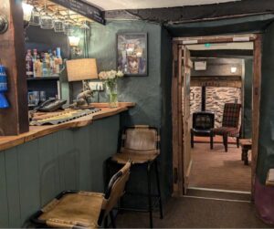 A cosy, rustic country in with a bar and chairs to the left and comfy chairs and wood burner through an open doorway.