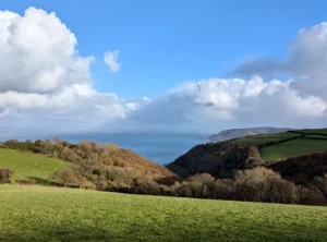 Undulating coastal landscape with green fields rolling into the distance with the sea appearing in the background under a blue sky.