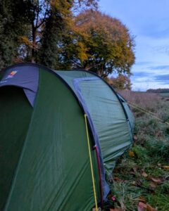 A tent pitched in the autumnal countryside countryside with a field to the right and trees to the left.