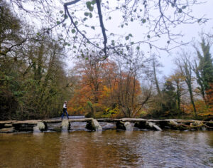 A lone walker crosses an ancient stone clapper bridge in Exmoor with autumnal trees in the background.