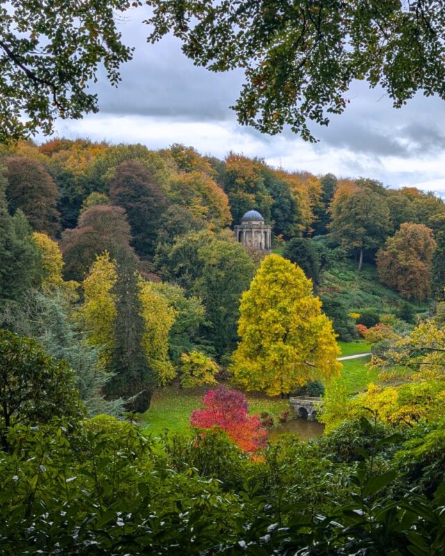 A wooded hillside with trees in full autumnal colours. Nestled in the trees is a replica Greek Pantheon