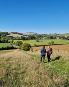 Two walkers are standing in a field with a map in hand. They are both looking out into the countryside in front of them