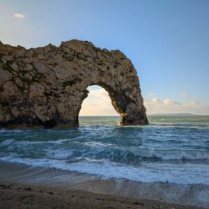A limestone arch reaching out into the sea with the waves hitting the shingle beach.
