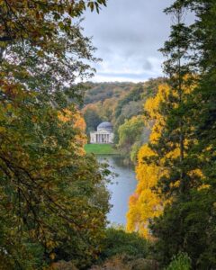 Scene of a lake surrounde by golden autumnal trees and a replica Greek pantheon on a hill in the distance