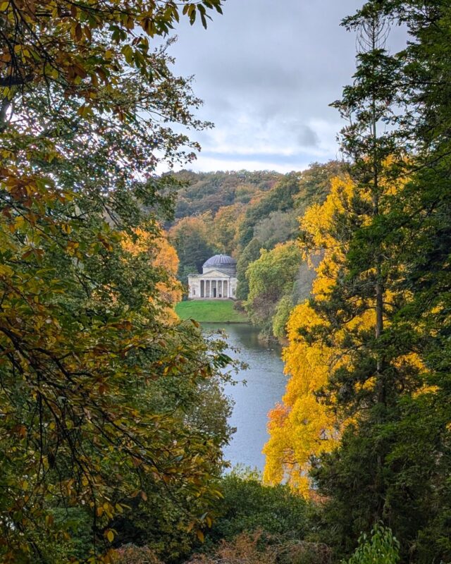 Scene of a lake surrounde by golden autumnal trees and a replica Greek pantheon on a hill in the distance