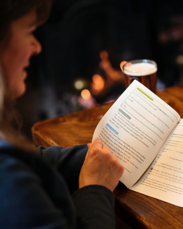 A woman is sat at a table in a country inn reading step-by-step walking instructions in a trail guide.