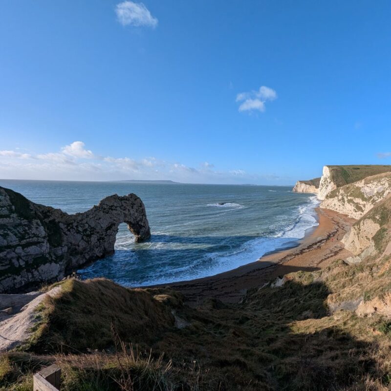 The iconic limestone arch Durdle Door taken from a clifftop, coastal cliffs stretching off to the right, the blue sea to the left