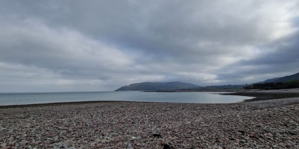 View of a grey pebble beach looking out onto the sea with the land curving around to the right.