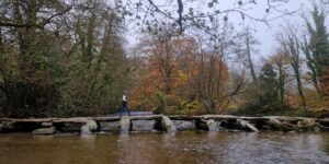 A figure walking across an ancient stone clapper bridge over a river with autumnal trees in the background.