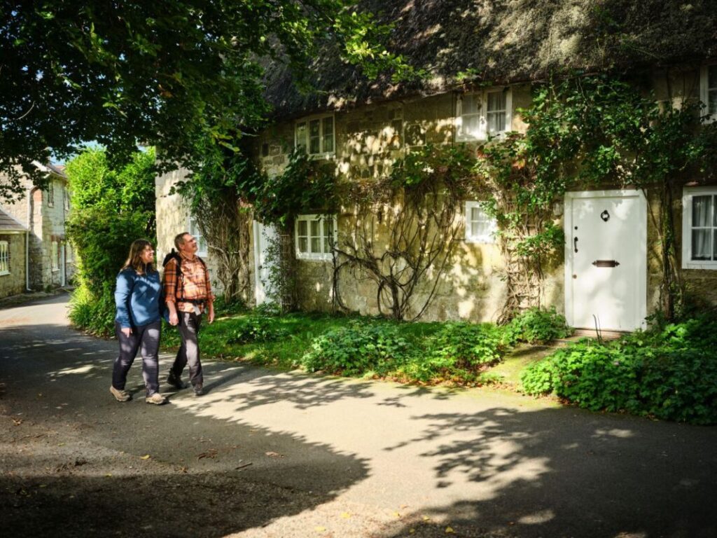 Two walkers walk past a traditional cottage in dappled sunlight.