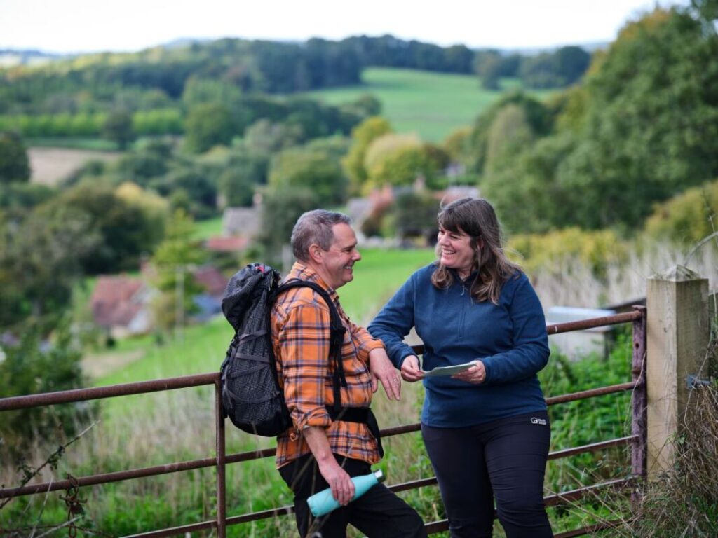 Two walkers lean on a farm gate with fields stretching off into the distance.