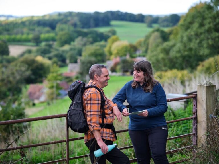 Two walkers lean on a farm gate with fields stretching off into the distance.